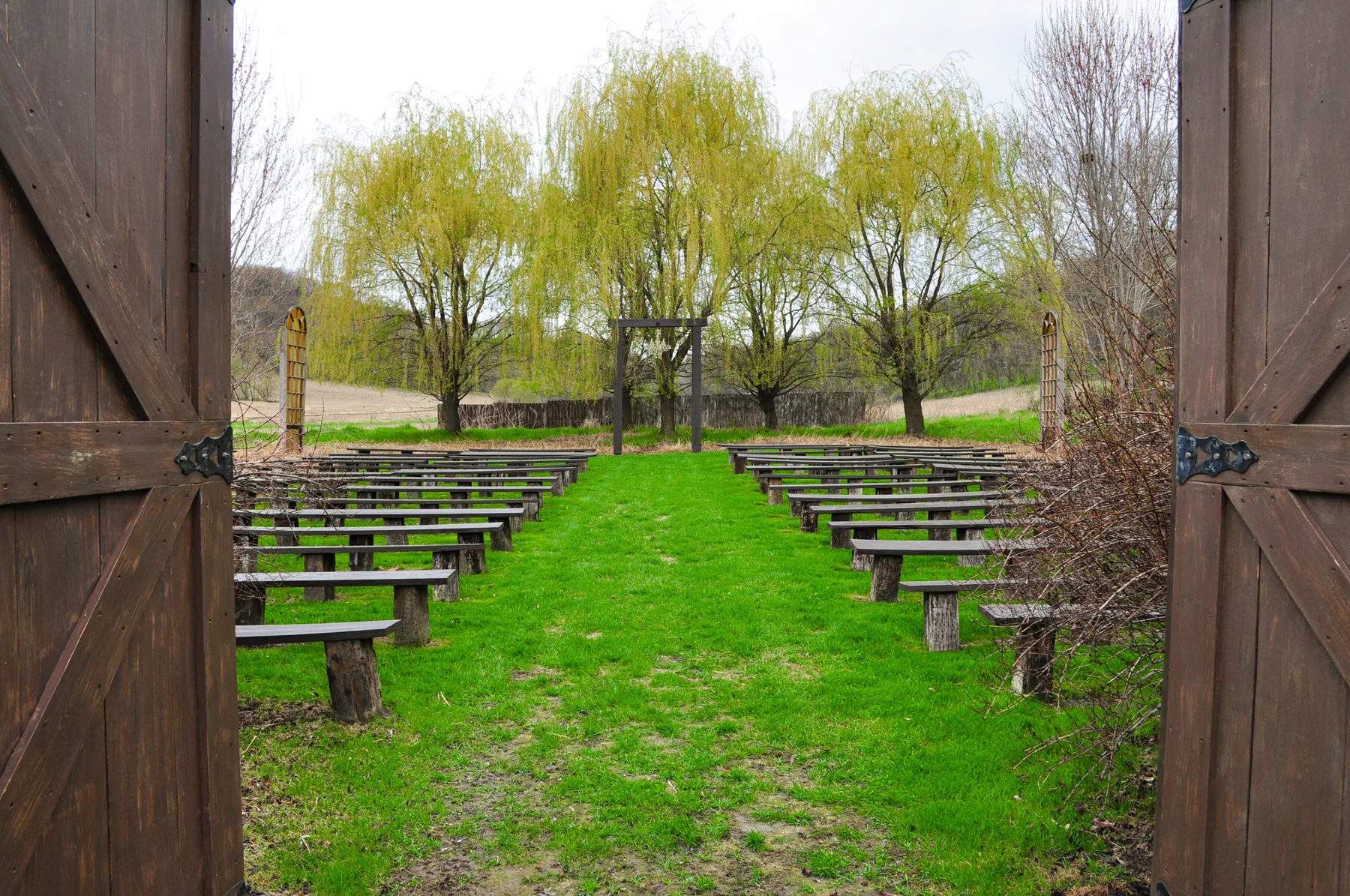 The view from the open barn doors out to rows of dark wooden benches and a simple wooden arch framed by weeping willows