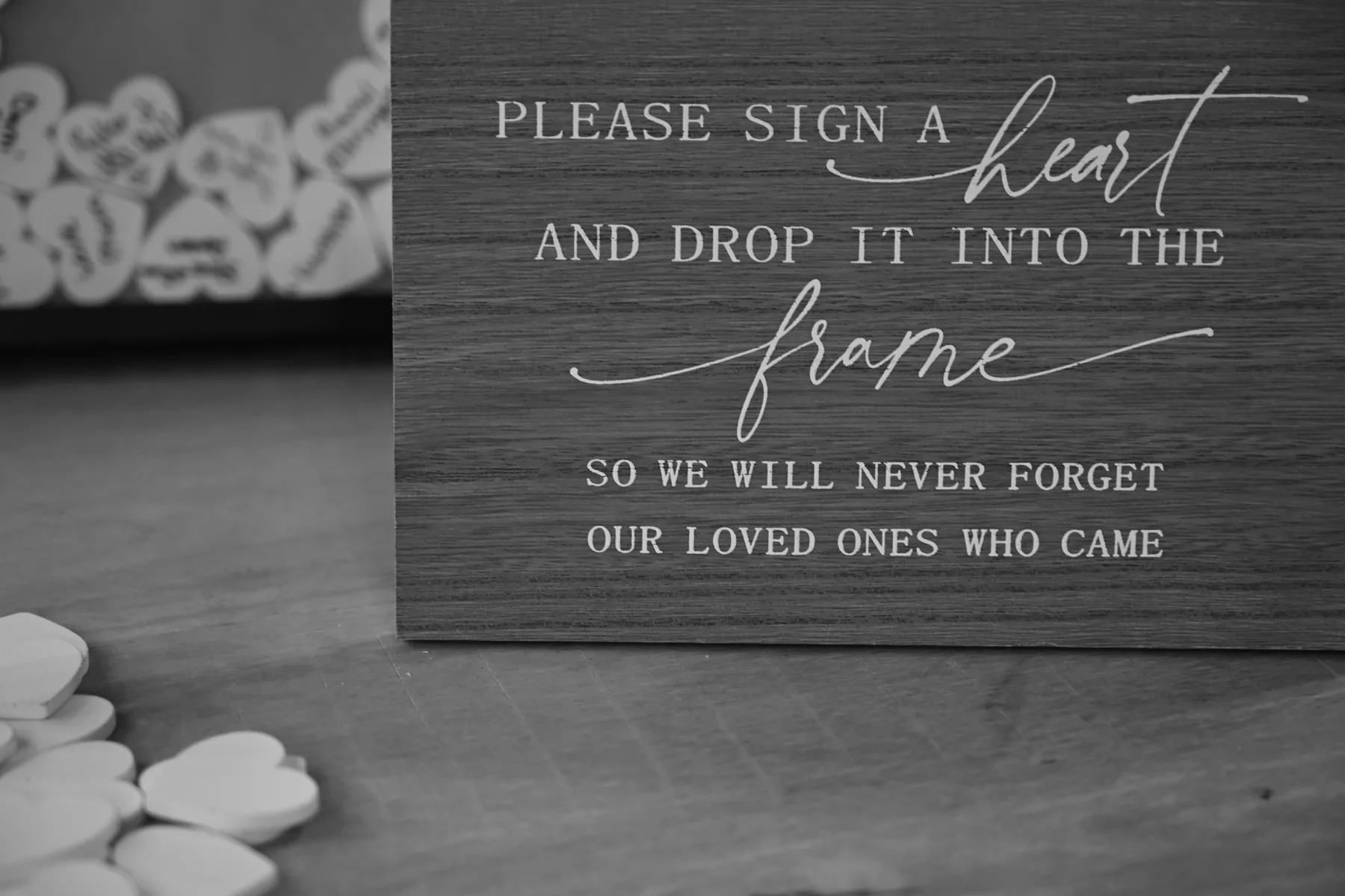 A small wooden guest book sign asking loved ones to sign a heart and drop it into the frame