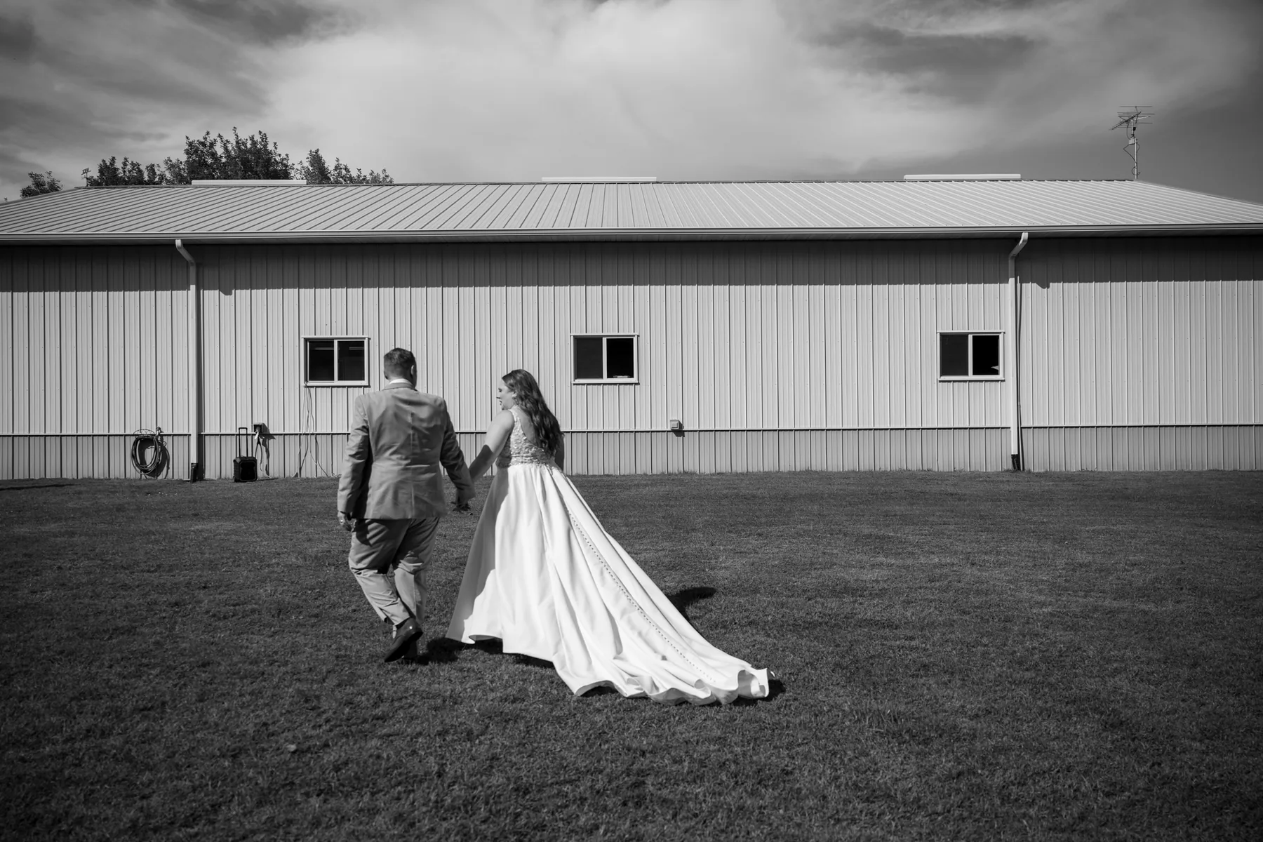 Kelsey and Michael walking hand in hand across the open lawn, her train trailing behind her and the pole barn stretched across the horizon