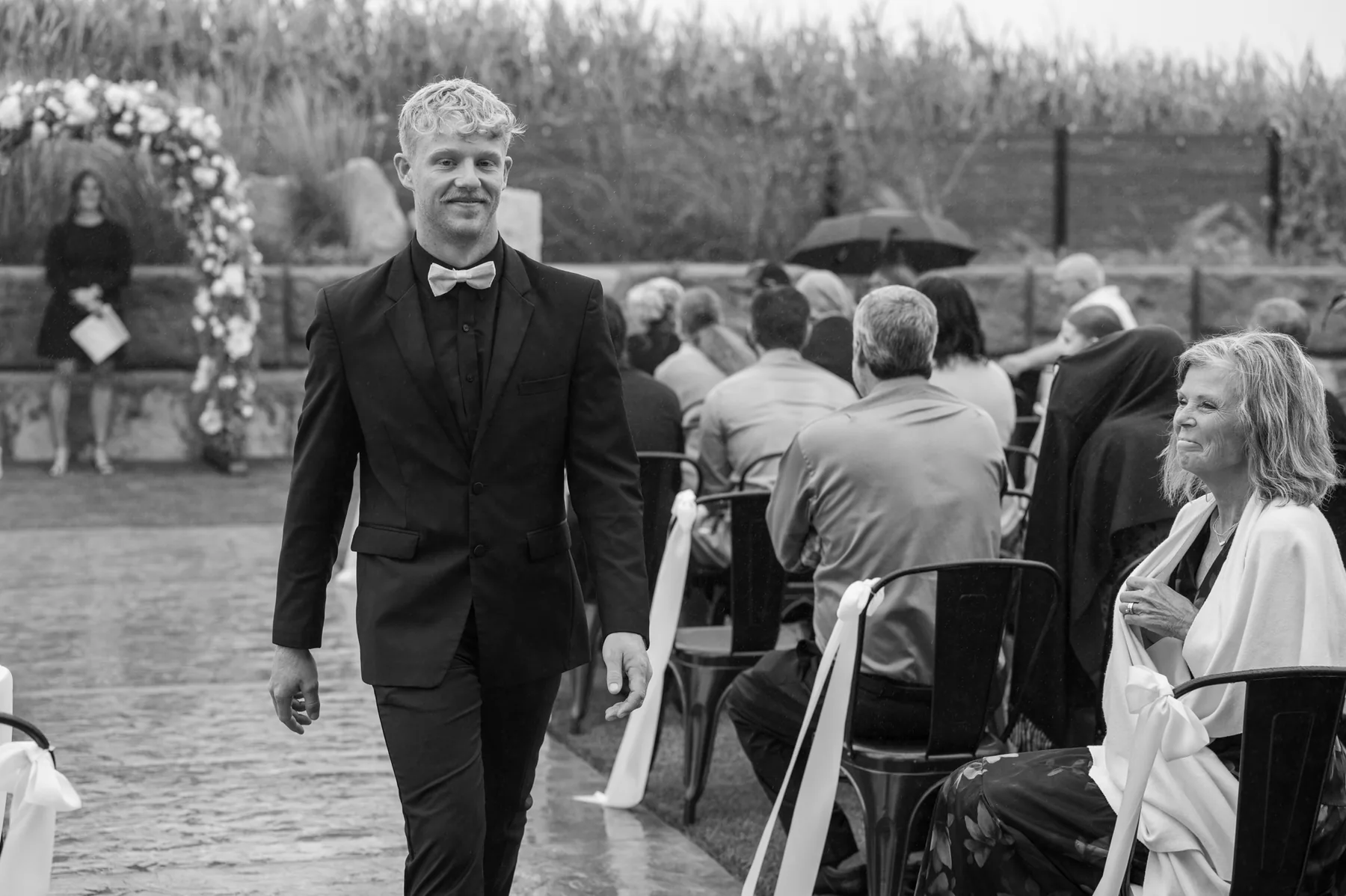 Gage walking the wet aisle in the rain, guests seated under umbrellas and the floral arch waiting ahead of him