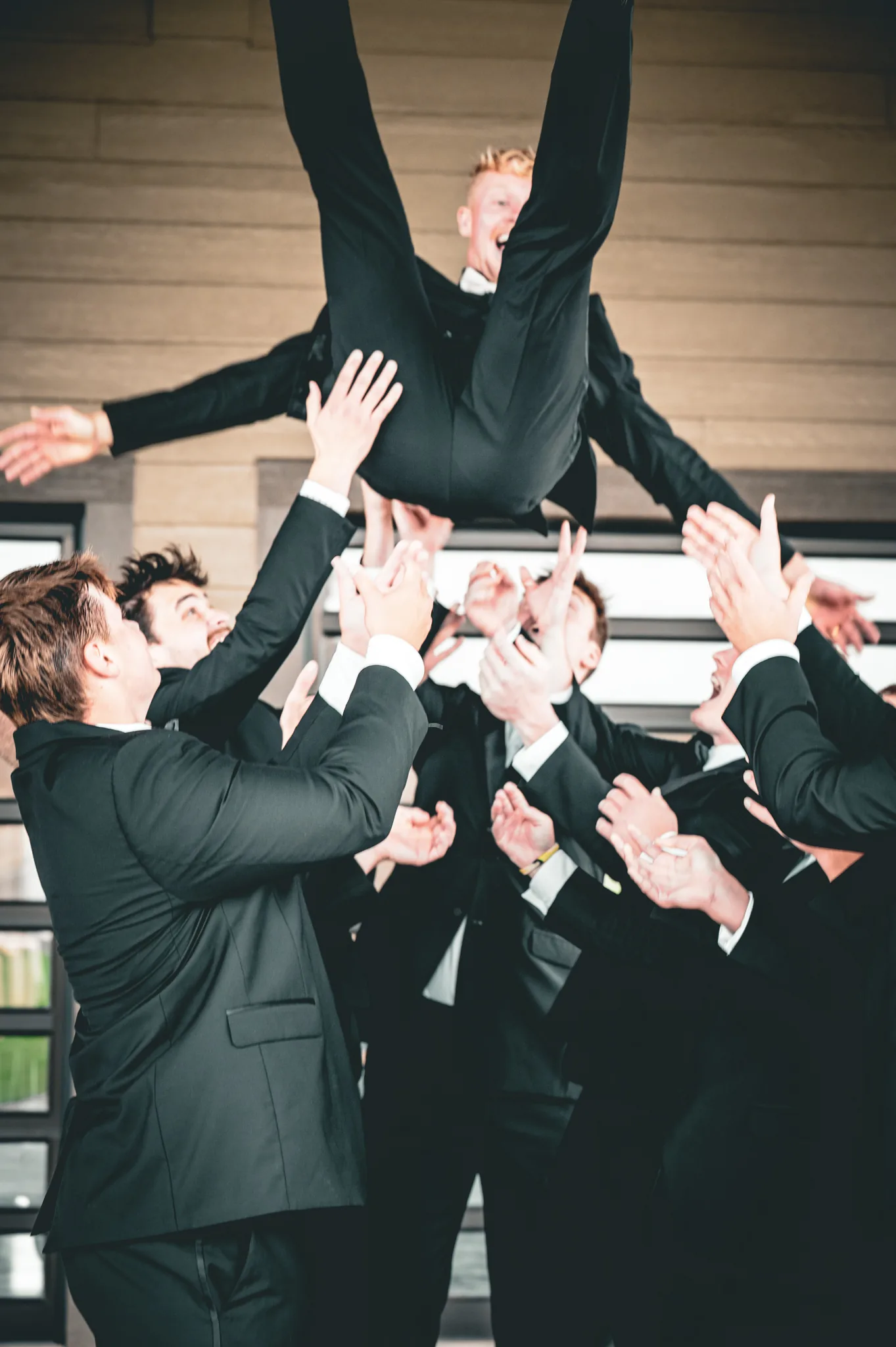 Groomsmen throwing one of the group into the air outside the venue, arms up and everyone mid-shout